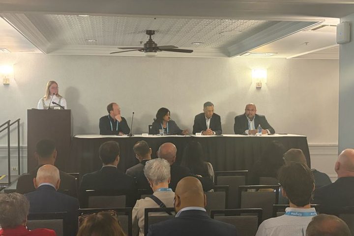 A conference panel on stage: a moderator stands at a podium on the left while four panelists in business attire sit at a long draped table with microphones, facing a seated audience wearing lanyards.