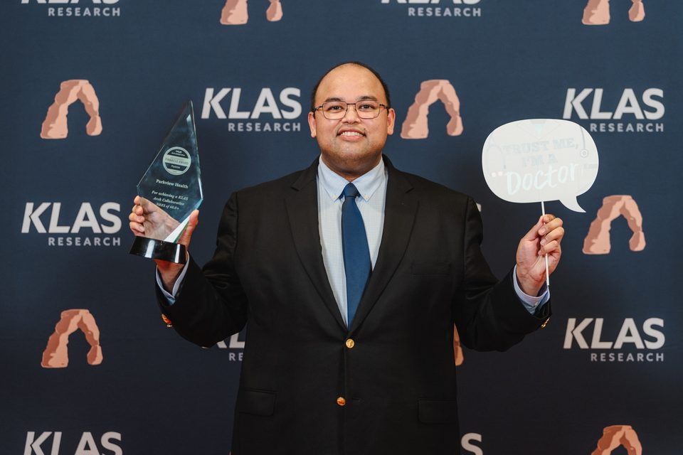 Hasan Ahmad smiling in front of a KLAS Research backdrop, holding a glass KLAS award trophy in one hand and a “Trust me, I’m a doctor” speech-bubble sign in the other.