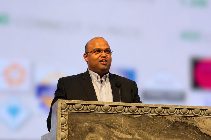 Hasan Ahmad in a dark suit jacket speaking at an ornately carved marble podium, with a blurred presentation screen behind him.